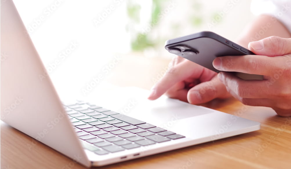 ノートパソコンを操作しながら片手でスマートフォンを持つ人物の手元の様子