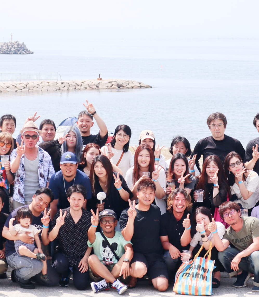 海辺で大人数のグループが集合写真を撮っており、皆がピースサインで笑顔を見せている