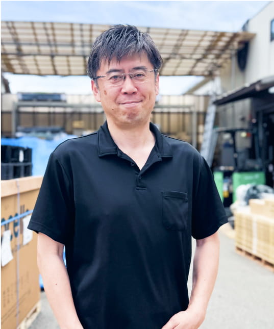 黒いポロシャツを着た男性が、屋外の資材置き場で微笑みながら立っている