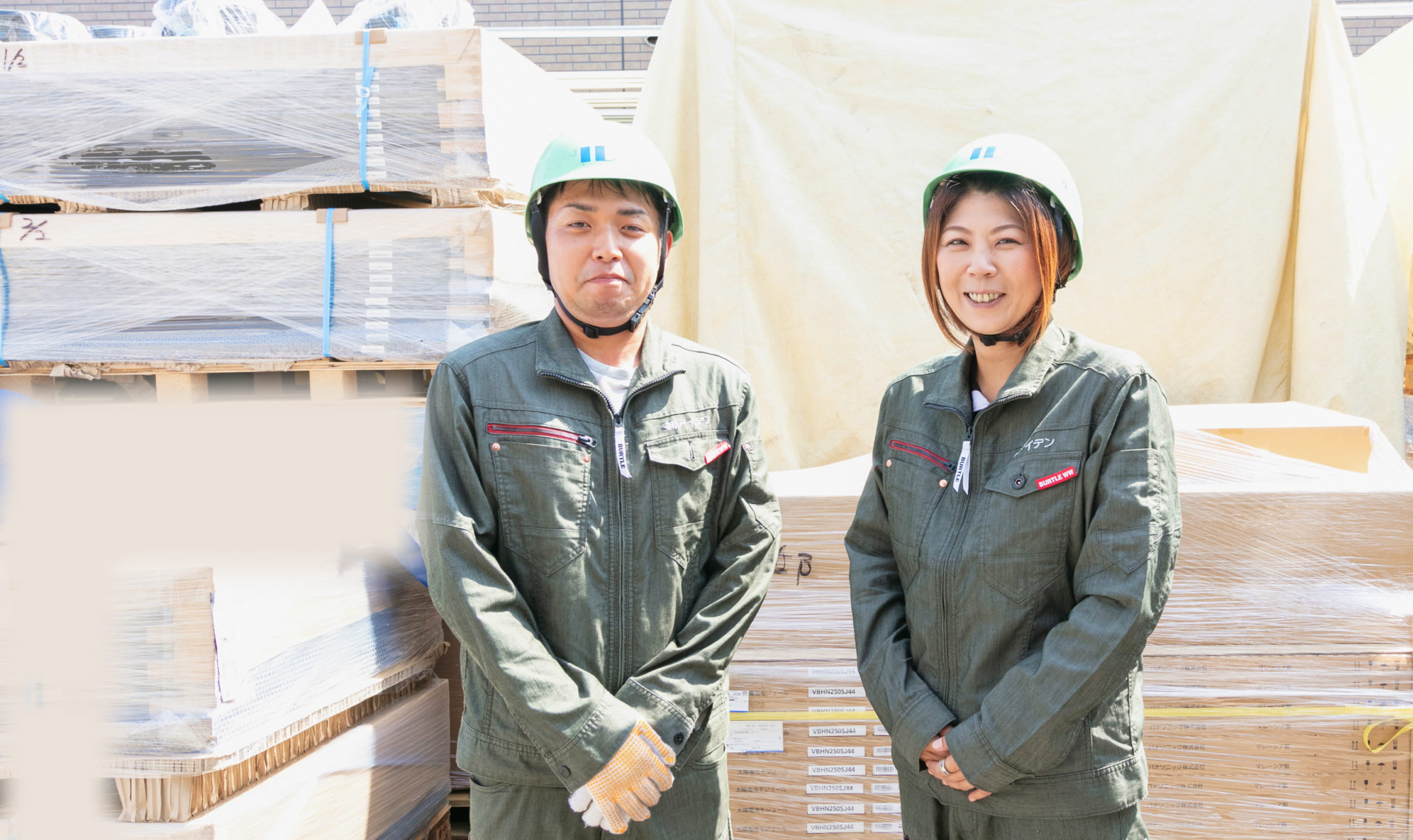 作業服とヘルメットを着用した男女2人が、屋外の資材置き場で並んで立っている様子