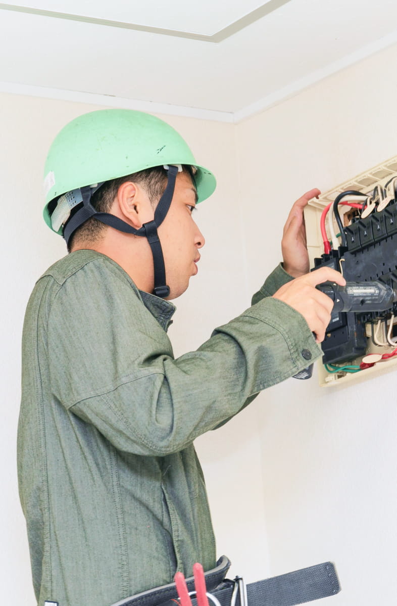 作業服とヘルメットを着用した男性が、室内で配電盤の点検作業を行っている