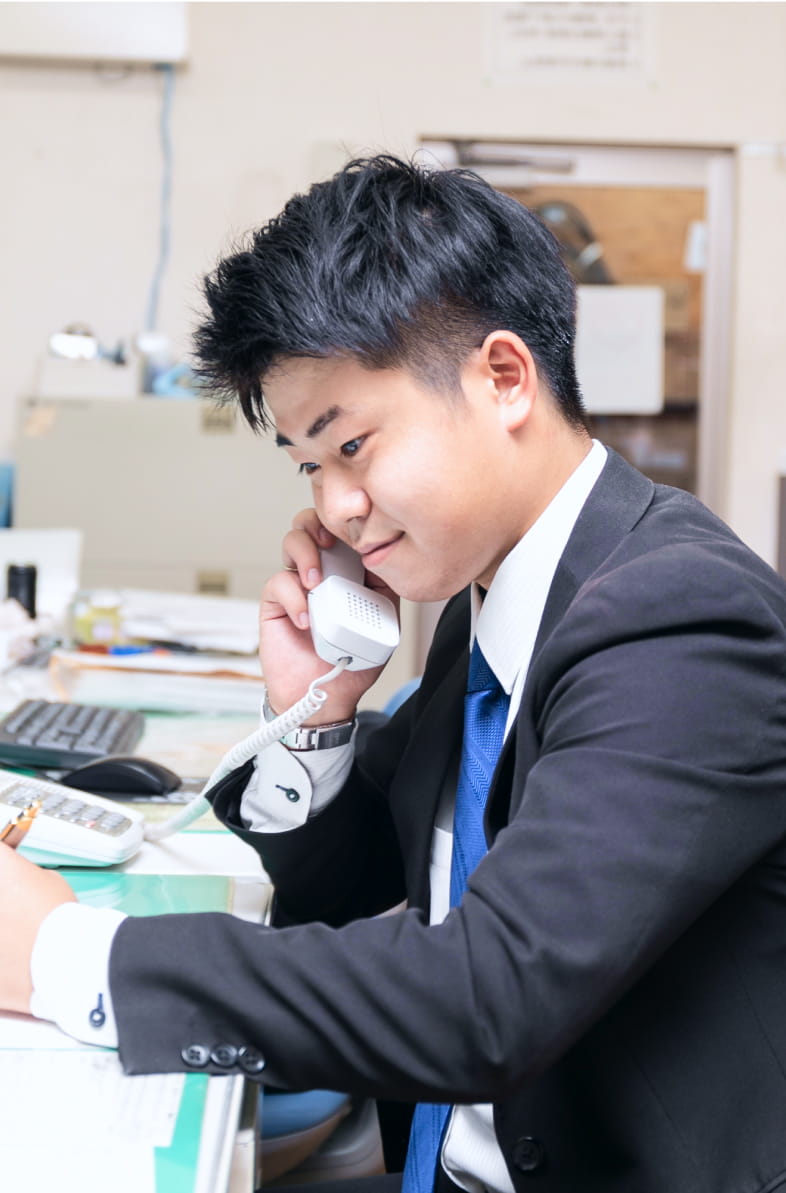 スーツ姿の若い男性がデスクで電話対応をしながらメモを取っている